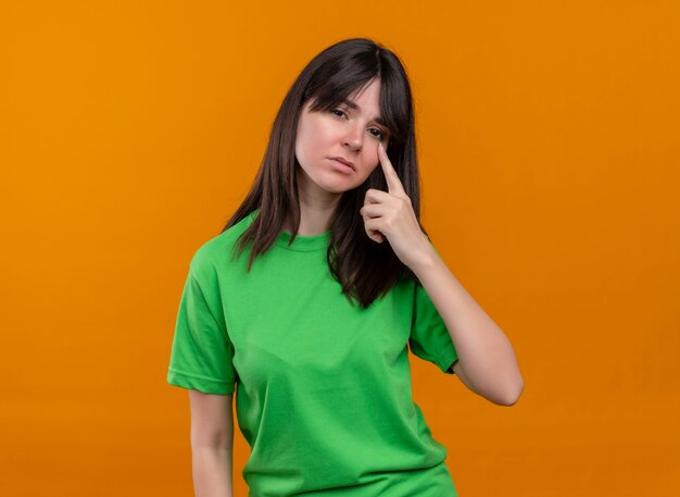 緑のシャツを着た悲しい若い白人の女の子が顔に手を置き、孤立したオレンジ色の背景でカメラを見る