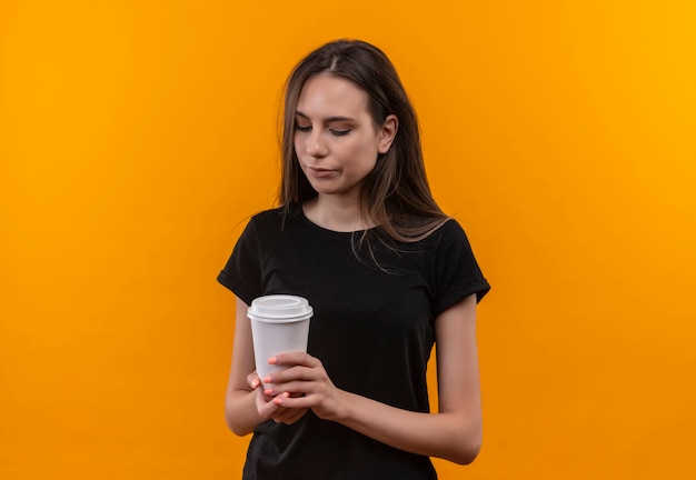 Грустная молодая кавказская девушка в черной футболке смотрит на чашку кофе на изолированном оранжевом фоне