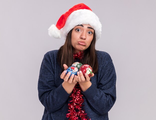 Sad young caucasian girl with santa hat and garland around neck holds glass ball ornaments 