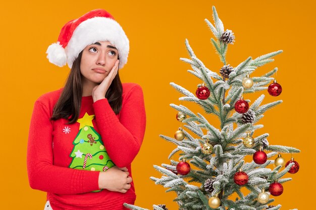 Sad young caucasian girl with santa hat puts hand on face and looks at side 