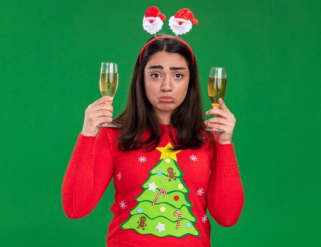 Sad young caucasian girl with santa headband holds glasses of champagne isolated on green background with copy space