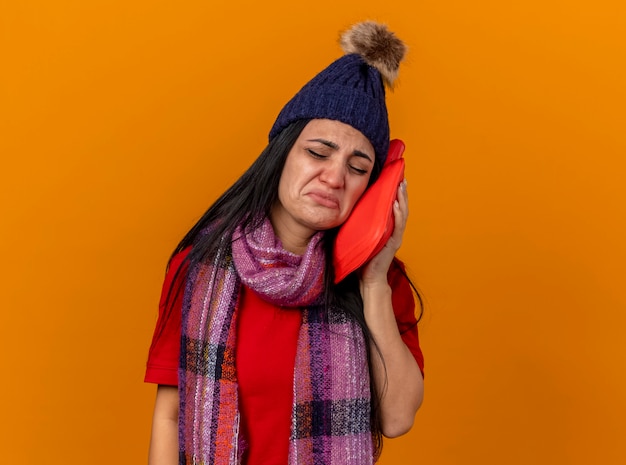 Sad young caucasian ill girl wearing winter hat and scarf touching face with hot water bag with closed eyes isolated on orange wall with copy space