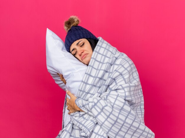 Sad young caucasian ill girl wearing winter hat and scarf wrapped in plaid hugging pillow putting head on it with closed eyes isolated on crimson background with copy space