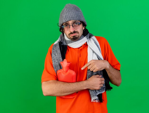 Грустный молодой кавказский больной мужчина в очках, зимняя шапка и шарф, держа и указывая на сумку с горячей водой, глядя в камеру, изолированную на зеленом фоне с копией пространства