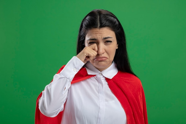 Sad young caucasian superhero girl crying and wiping her tears isolated on green wall with copy space