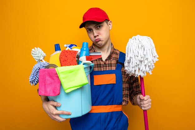 Sad young cleaning guy wearing uniform and cap holding bucket of cleaning tools with mop 