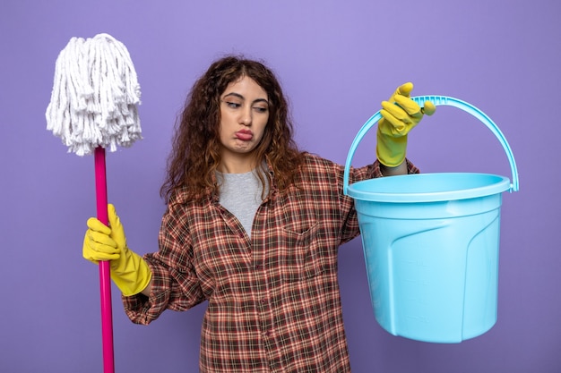 Sad young cleaning woman wearing gloves holding mop looking at bucket in her hand 