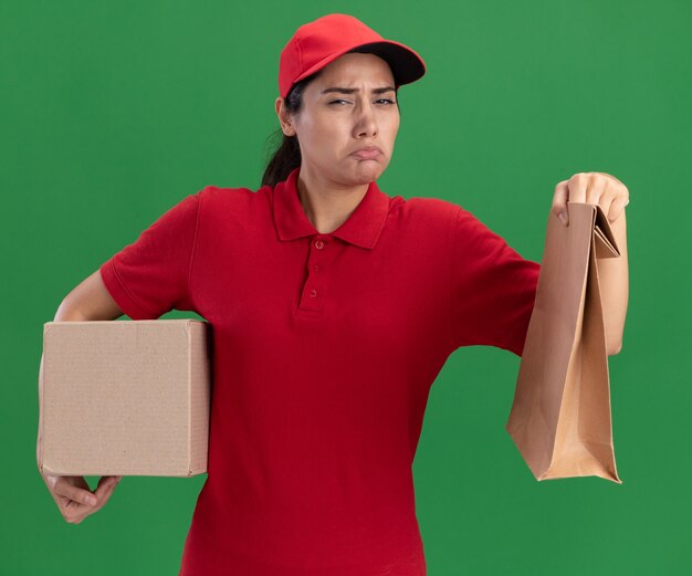 Sad young delivery girl wearing uniform and cap holding box with paper food package isolated on green wall