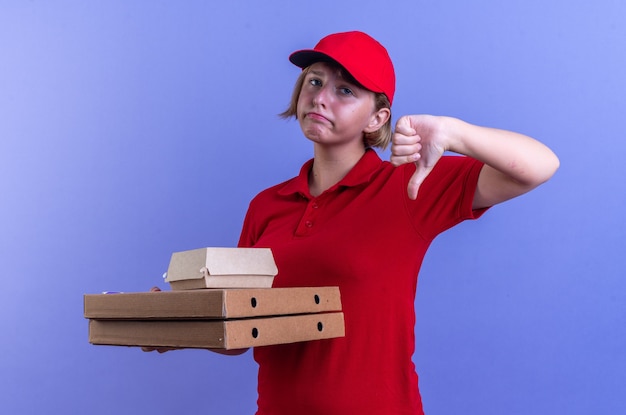 грустная молодая доставщица в униформе и кепке держит бумажную коробку для еды на коробках для пиццы, показывая большой палец вниз, изолированную на синей стене