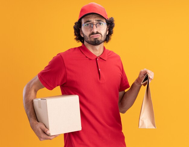 オレンジ色の壁で隔離の正面を見てカードボックスと紙のパッケージを保持している眼鏡をかけている赤い制服と帽子の悲しい若い配達人
