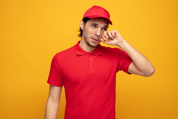 sad young delivery man wearing uniform and cap looking at side crying wiping eye with hand isolated on yellow background