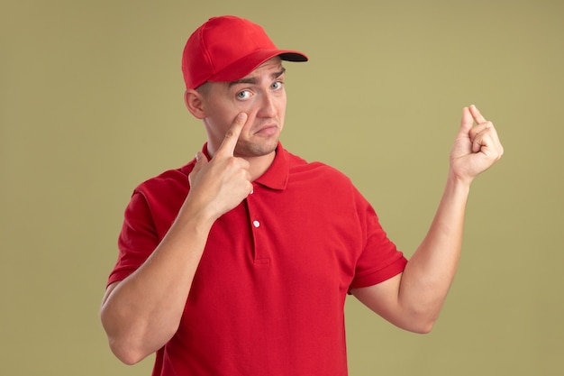 Sad young delivery man wearing uniform and cap pulling down eye lids showing tip gesture isolated on olive green wall