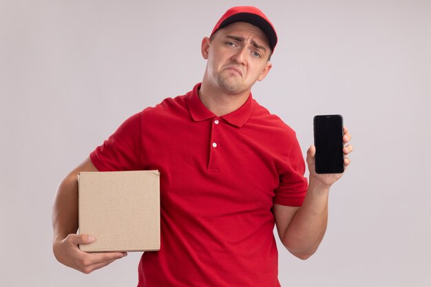 Sad young delivery man wearing uniform with cap holding box and phone isolated on white wall