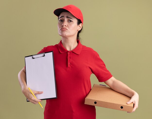 Triste giovane donna delle consegne in uniforme e berretto che tiene un pacchetto di pizza che mostra appunti con la matita in mano guardando la parte anteriore isolata sul muro verde oliva