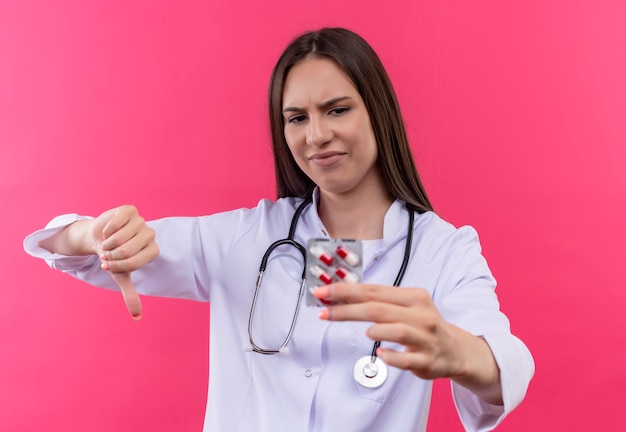 Грустная молодая девушка-врач в медицинском халате со стетоскопом смотрит на таблетки на руке, опустив большой палец на изолированный розовый фон