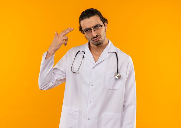 Грустный молодой врач в медицинских очках в медицинском халате со стетоскопом показывает жест пистолета на изолированной желтой стене с копией пространства