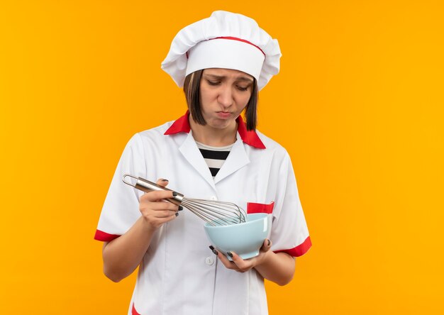 Грустная молодая женщина-повар в униформе шеф-повара держит и смотрит на венчик и миску, изолированные на оранжевой стене