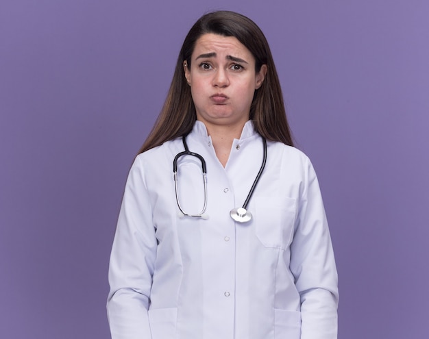 Sad young female doctor wearing medical robe with stethoscope blowing cheeks isolated on purple wall with copy space