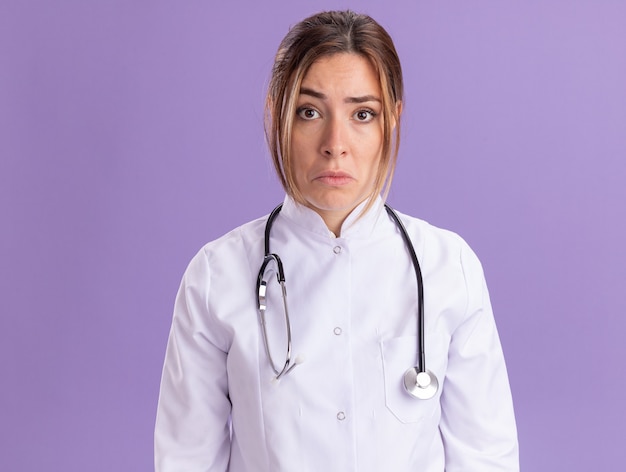 Грустная молодая женщина-врач в медицинском халате со стетоскопом, изолированным на фиолетовой стене