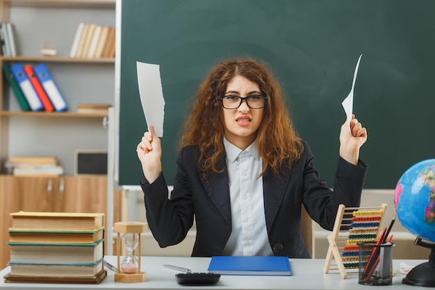 Free photo sad young female teacher wearing glasses tear paper sitting at desk with school tools in classroom