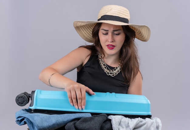 Sad young female traveler wearing black undershirt in hat holding open suitcase on white wall