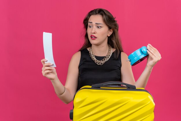 Sad young female traveler wearing black undershirt holding alarm clock and ticket looking at ticket on red wall