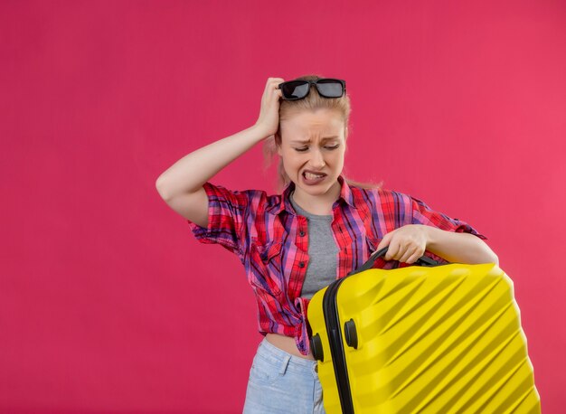 Sad young female traveler wearing red shirt in glasses holding suitcase put her hand on head on isolated pink wall