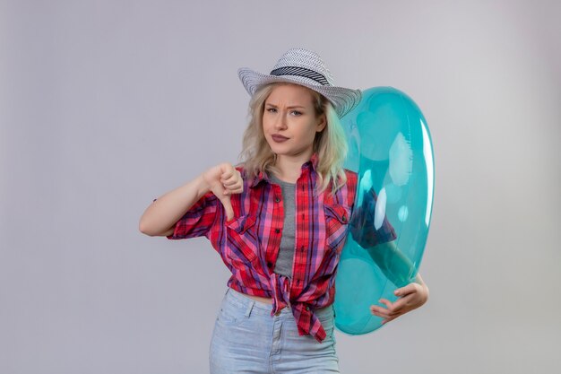 Sad young female traveler wearing red shirt in hat holding inflatable ring her thumb down on isolated white wall