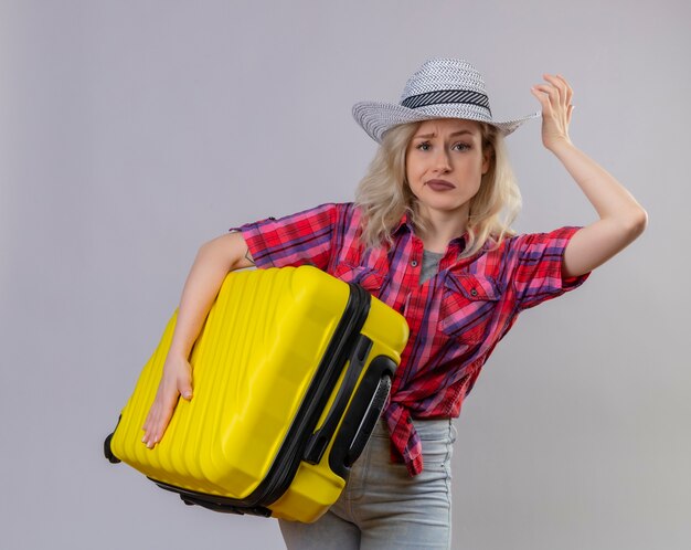 Sad young female traveler wearing red shirt in hat holding suitcase on isolated white wall