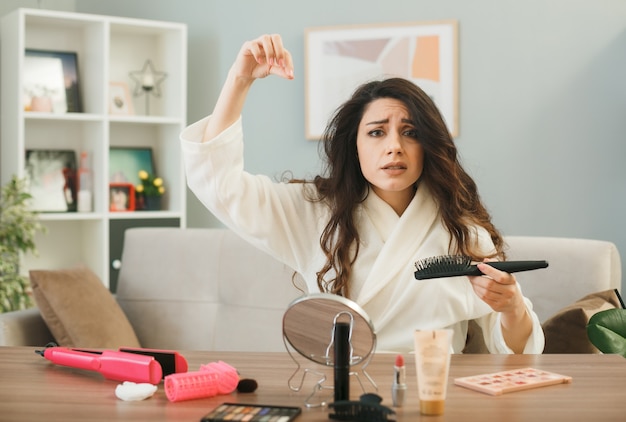 Sad young girl holding comb sitting at table with makeup tools in living room