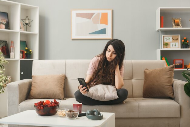Sad young girl holding and looking at phone sitting on sofa behind coffee table in living room