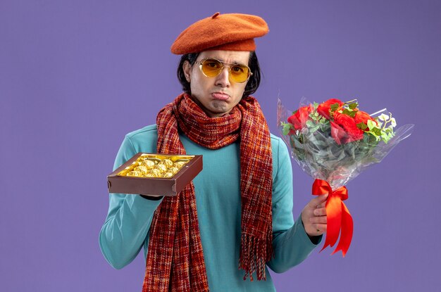 Sad young guy on valentines day wearing hat with scarf and glasses holding bouquet with box of candies isolated on blue background