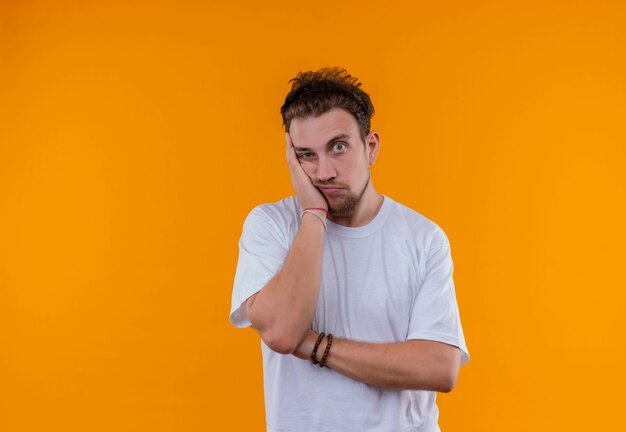 Sad young guy wearing white t-shirt put his hand on cheek on isolated orange background