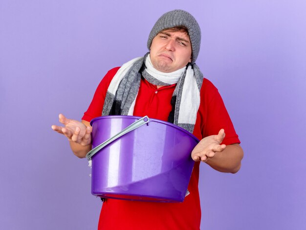 Sad young handsome blonde ill man wearing winter hat and scarf holding plastic bucket  isolated on purple wall