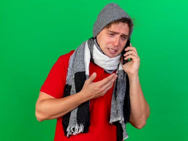 Free photo sad young handsome blonde ill man wearing winter hat and scarf talking on phone looking at side keeping hand in air isolated on green background with copy space