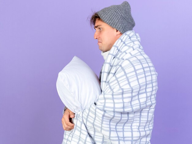 Sad young handsome blonde ill man wearing winter hat and scarf wrapped in plaid standing in profile view holding pillow looking straight isolated on purple background with copy space