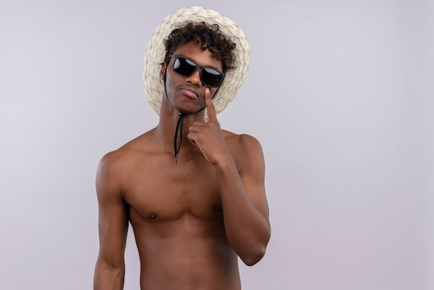 Free photo a sad young handsome dark-skinned man with curly hair wearing sun hat and sunglasses pointing with index finger on cheek while