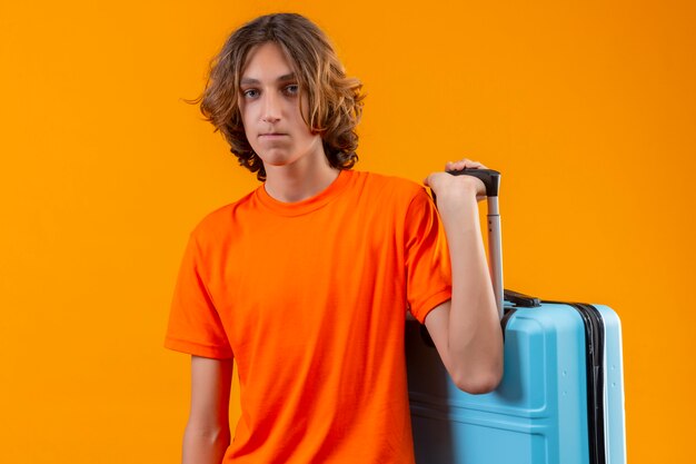 不幸な顔立ちでカメラを見て旅行スーツケースを保持しているオレンジ色のtシャツで悲しい若いハンサムな男
