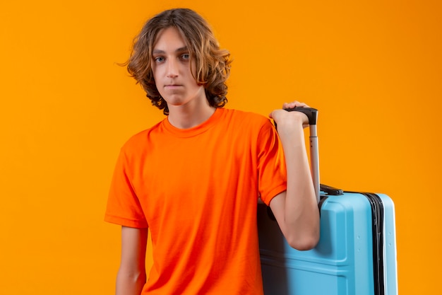 불행한 얼굴 서있는 카메라를 찾고 여행 가방을 들고 오렌지 티셔츠에 슬픈 젊은 잘 생긴 남자
