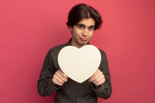 Sad young handsome guy wearing black t-shirt holding heart shape gesture isolated on pink wall