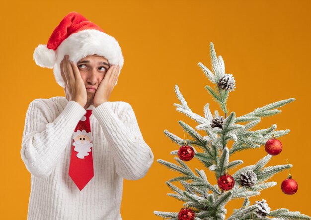 Грустный молодой красивый парень в новогодней шапке и галстуке санта-клауса стоит возле украшенной елки, держа руки на лице, глядя в сторону, изолированную на оранжевом фоне