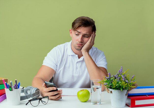 Sad young handsome male student sitting at desk with school tools holding and looking at phone isolated on olive green
