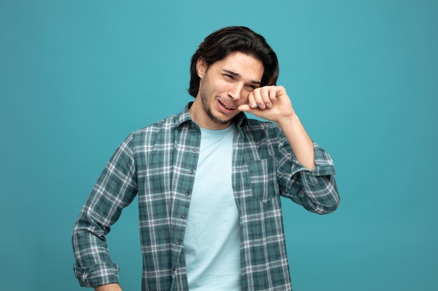 sad young handsome man looking at side wiping eye with hand crying isolated on blue background