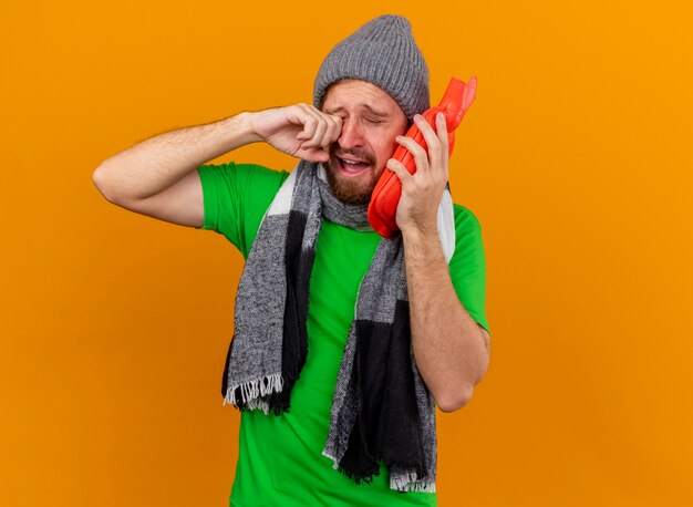 Sad young handsome slavic ill man wearing winter hat and scarf holding hot water bag touching face with it wiping tears isolated on orange background with copy space