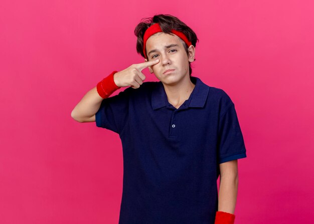 Sad young handsome sporty boy wearing headband and wristbands with dental braces looking at front pointing at his nose isolated on pink wall with copy space