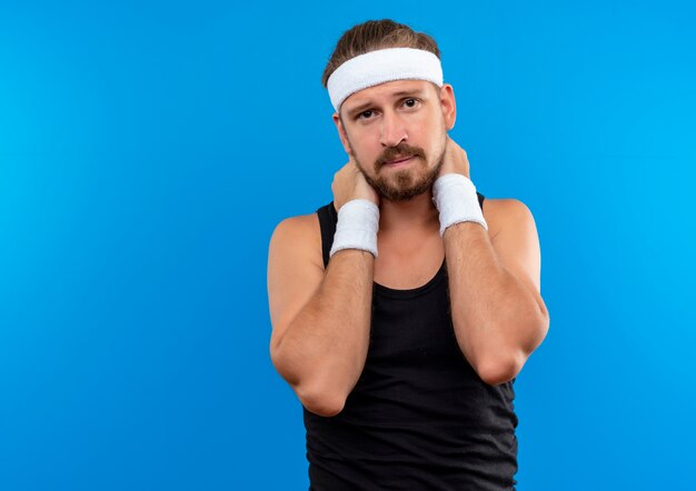 Sad young handsome sporty man wearing headband and wristbands putting hands on neck isolated on blue space 