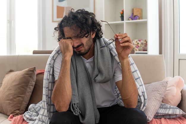 Free photo sad young ill caucasian man wrapped in plaid with scarf around his neck wipes his tears and holding optical glasses sitting on couch at living room