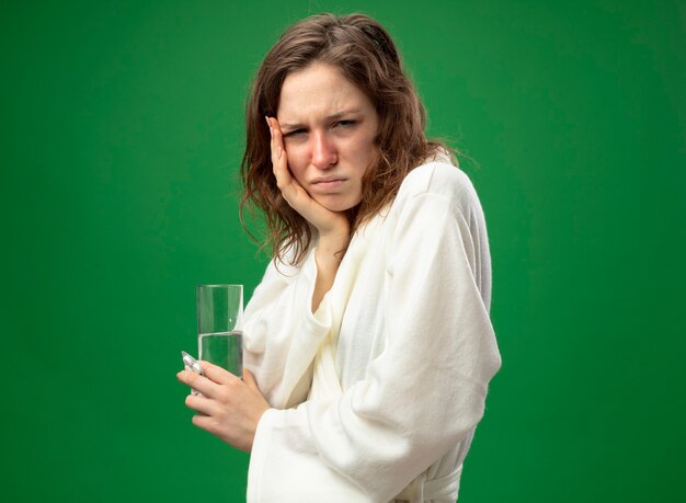 Sad young ill girl looking straight ahead wearing white robe holding glass of water putting hand on cheek isolated on green