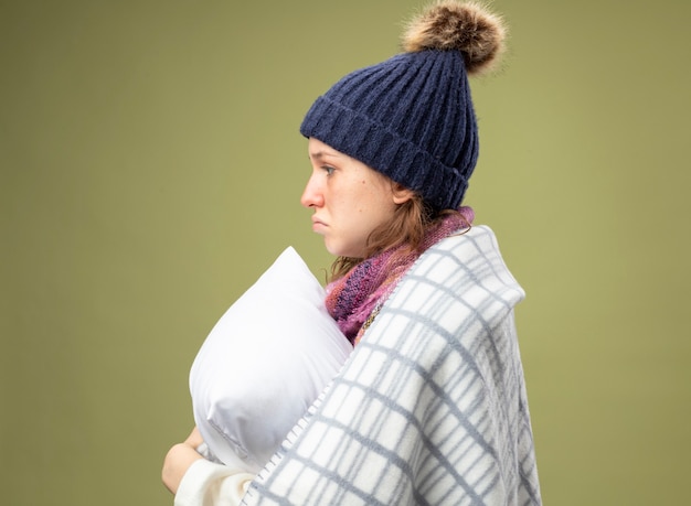 Triste giovane ragazza malata in piedi in vista di profilo che indossa una veste bianca e cappello invernale con sciarpa avvolta in un plaid abbracciato cuscino