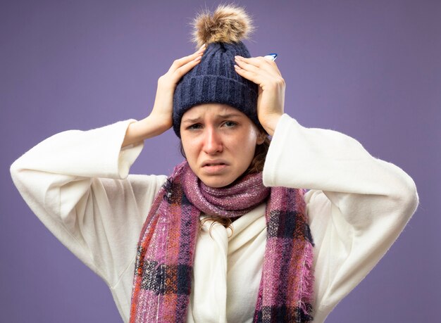 sad young ill girl wearing white robe and winter hat with scarf grabbed head isolated on purple